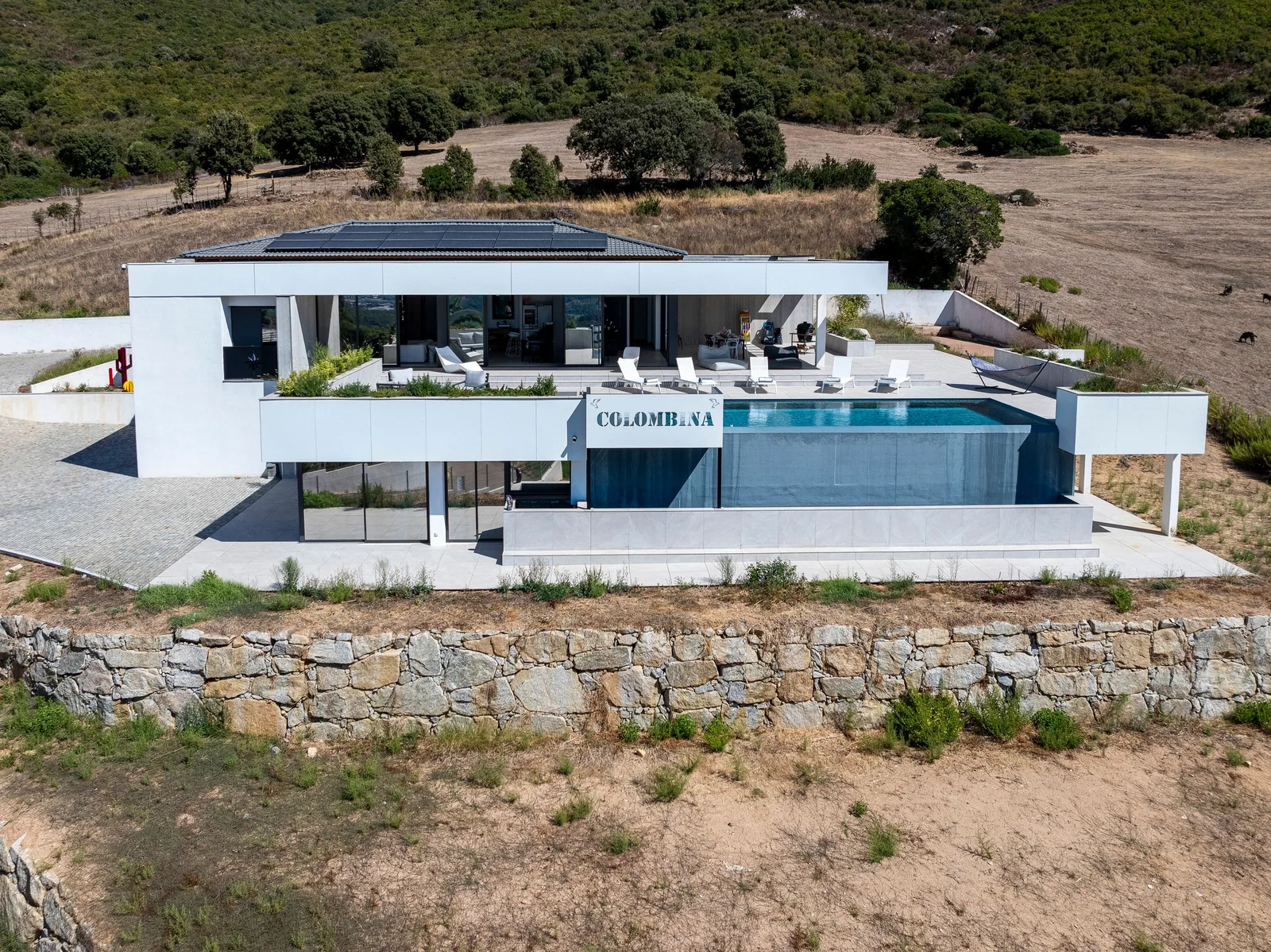 Façade de la villa au-dessus d'un muret de pierre sèche, piscine et terrasse face au maquis