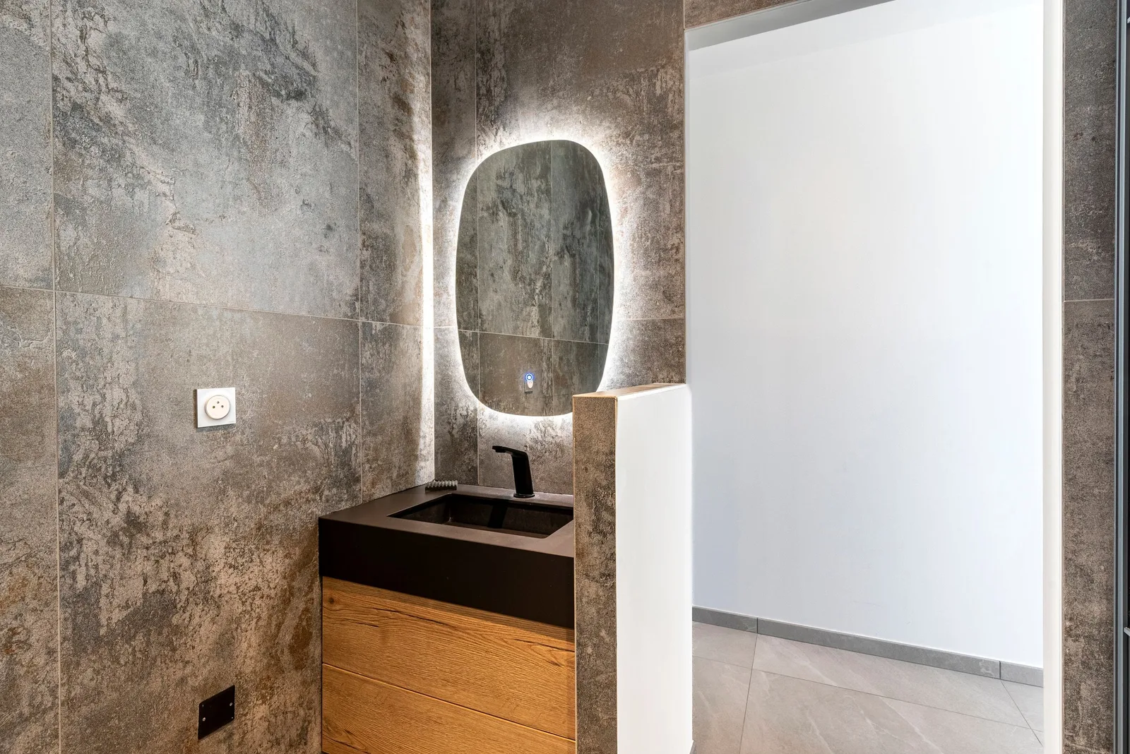 Vasque noire sur meuble en bois clair sous un miroir rétroéclairé, murs gris pierre