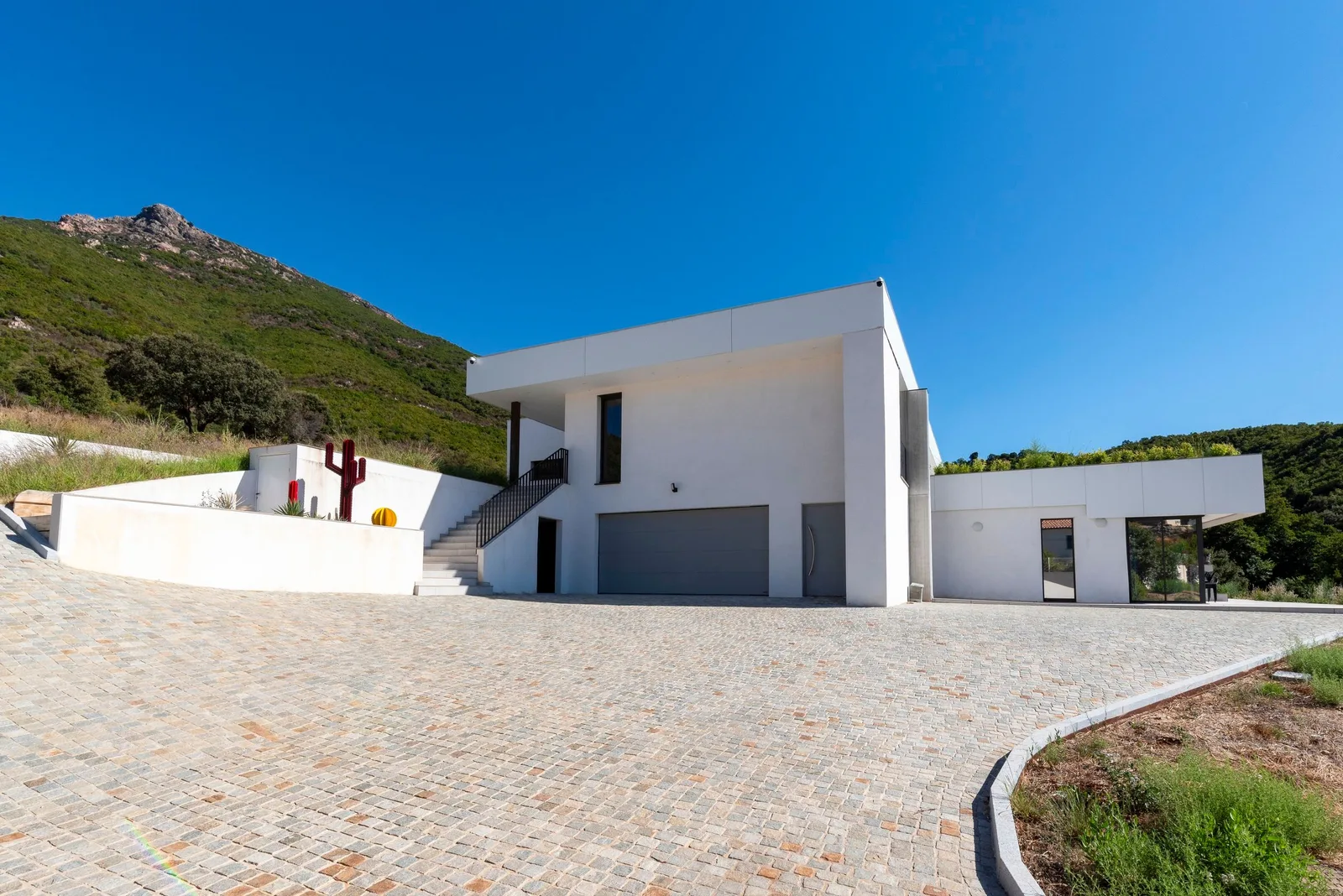 Façade d'entrée et garage de la villa sur une cour pavée, crête rocheuse et ciel bleu