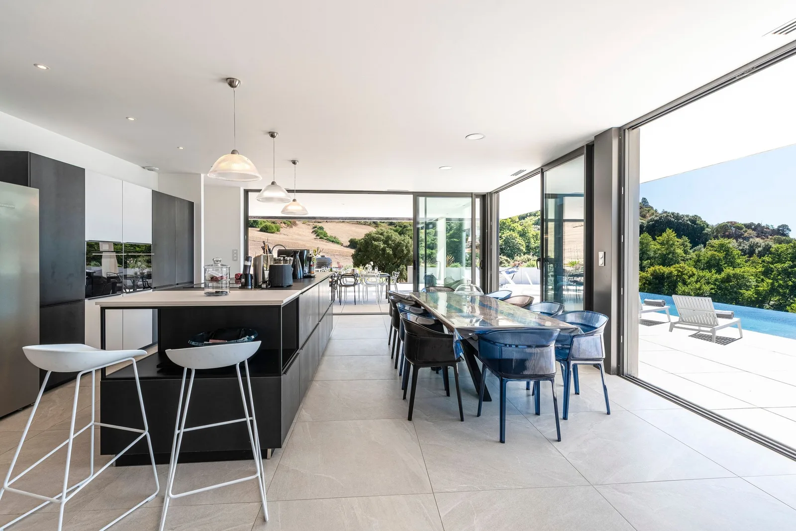 Îlot de cuisine avec tabourets blancs, salle à manger et baie ouverte sur la piscine