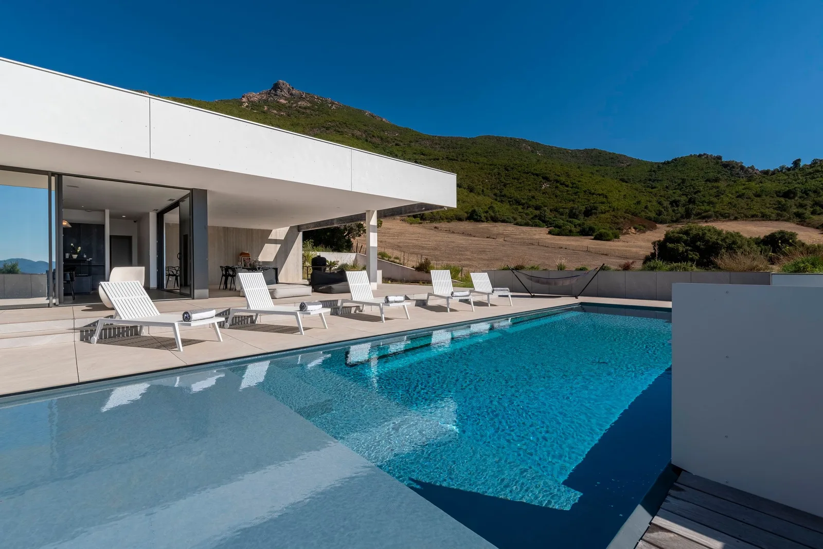 Piscine à débordement le long de la façade vitrée, bains de soleil et sommet rocheux au loin