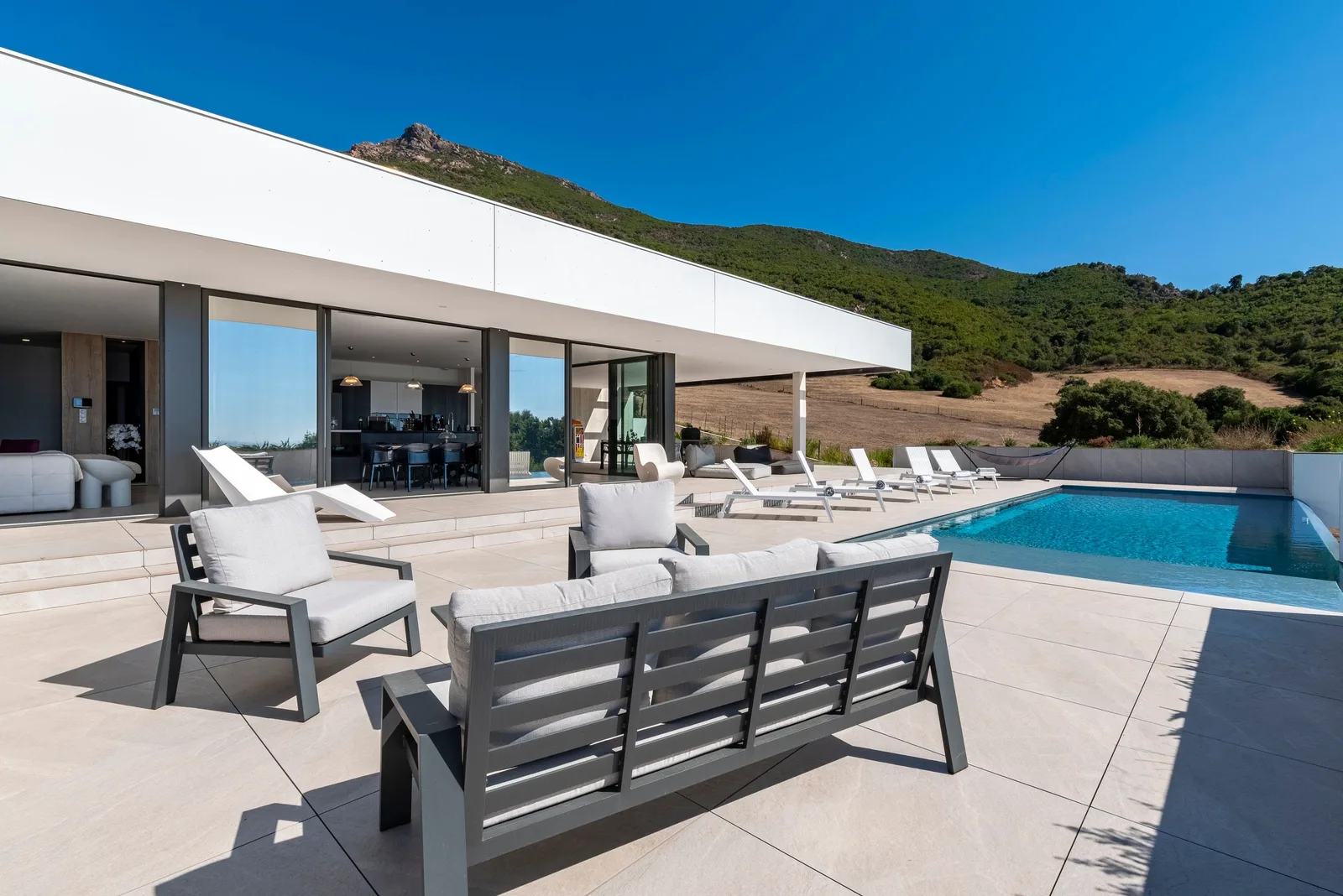 Salon extérieur en aluminium au bord de la piscine, coussins gris et crête rocheuse derrière la villa