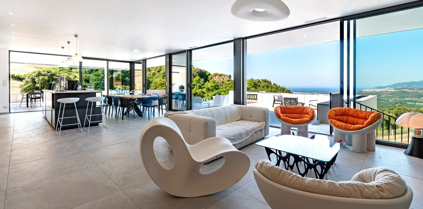 Séjour ouvert aux fauteuils orange et rocking-chair blanc face aux baies vitrées et à la mer