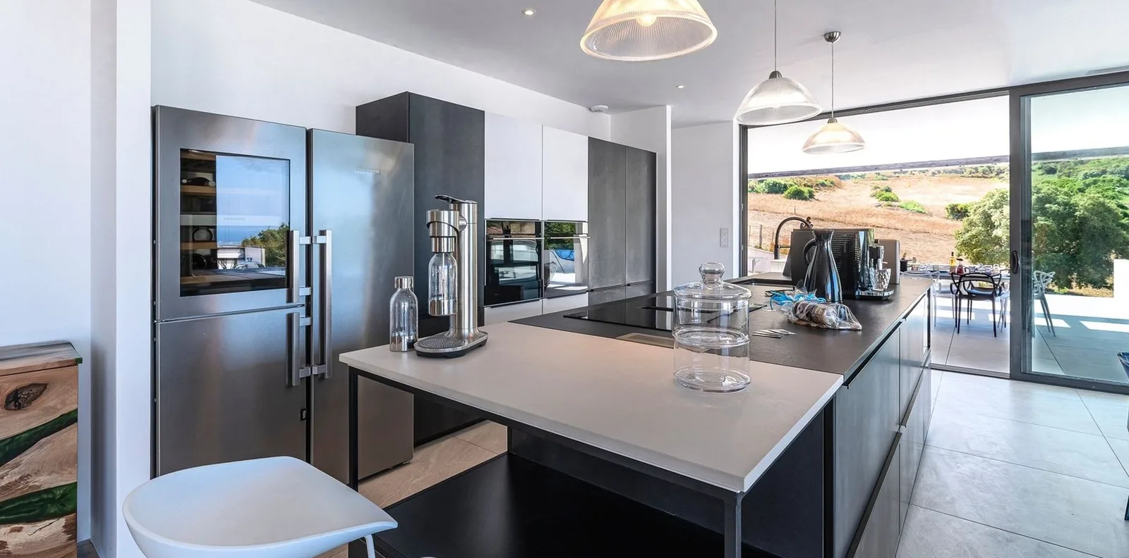 Cuisine contemporaine avec îlot central, électroménager inox et grande baie ouverte sur les collines