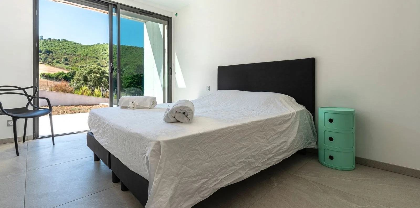 Chambre lumineuse ouverte sur le maquis par une baie vitrée, chevet vert et fauteuil design noir