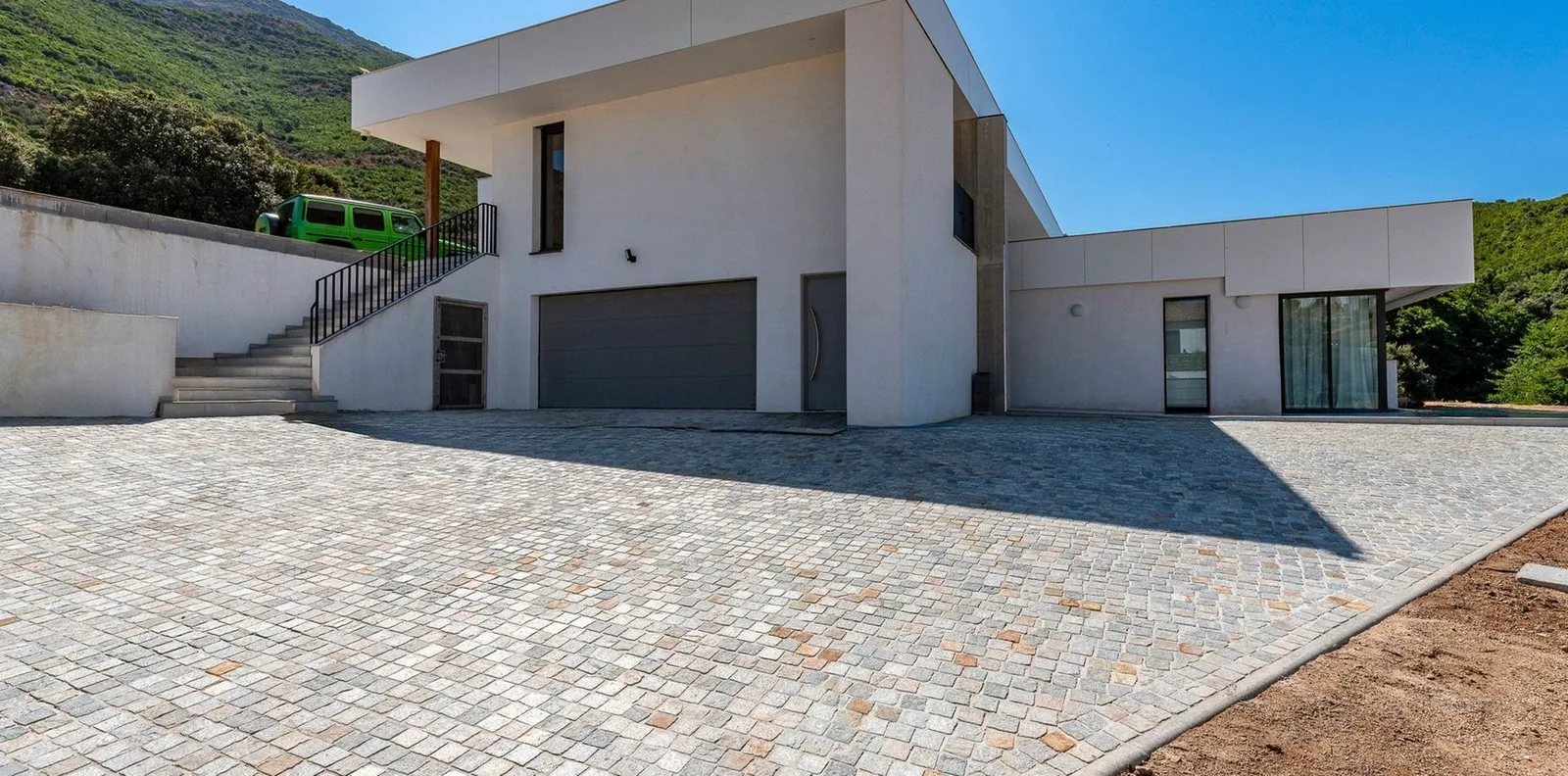 Façade et garage de la villa desservis par une vaste cour pavée, adossés à la colline verdoyante