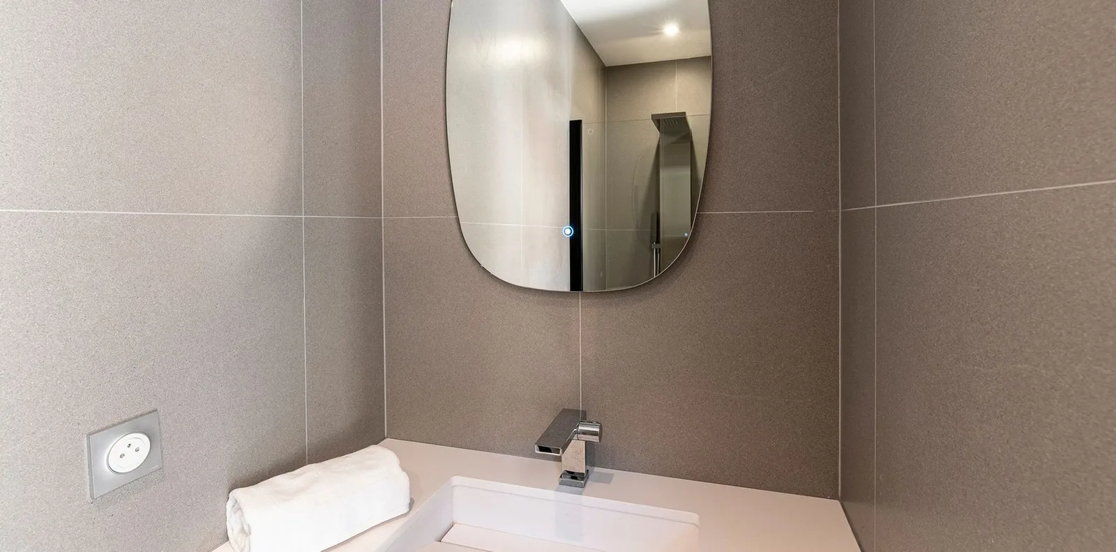Salle de bain taupe minimaliste, miroir tactile ovale et plan vasque blanc avec robinetterie chromée