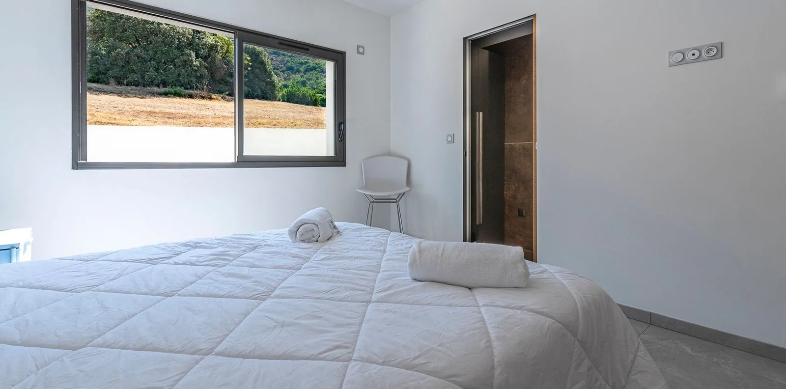 Chambre blanche avec grand lit, chaise filaire et salle d'eau attenante ouverte sur le maquis