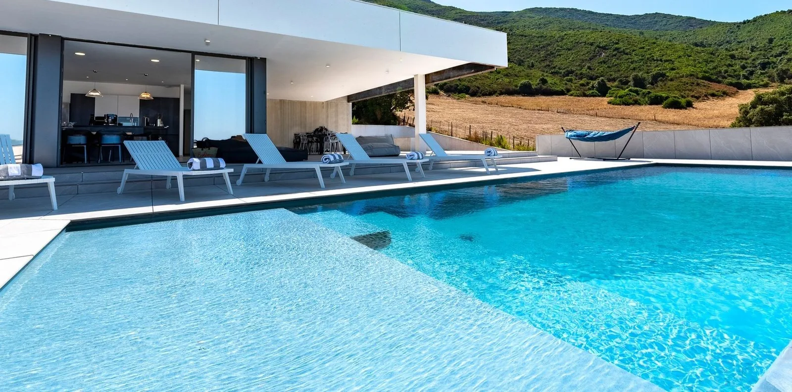 Piscine turquoise bordée de transats blancs et d'un hamac face aux collines boisées