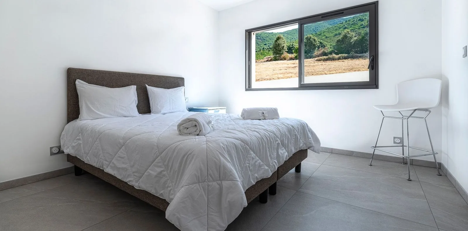 Chambre épurée au lit double blanc et fenêtre cadrant les champs corses
