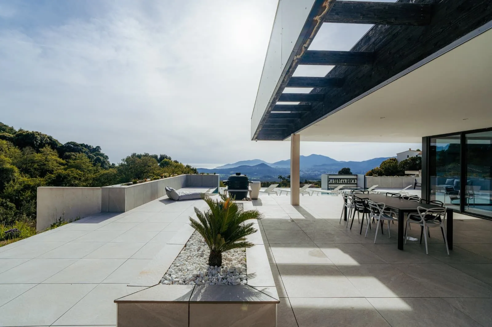 Vaste terrasse sous pergola en bois avec table à manger, barbecue et jardinière de palmiers